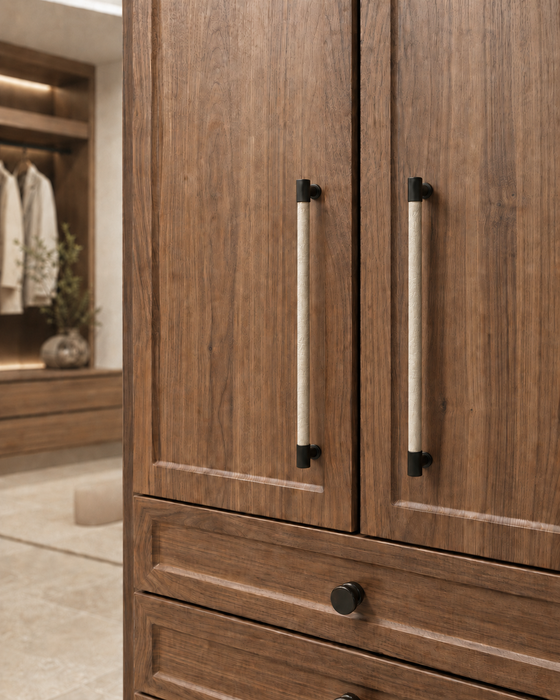 Close-up of a wooden cabinet with two doors and two long vertical handles, plus a drawer with a round knob below. The cabinet is shown in a real interior setting with a softly blurred background.