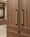 Close-up of a wooden cabinet with two doors and two long vertical handles, plus a drawer with a round knob below. The cabinet is shown in a real interior setting with a softly blurred background.