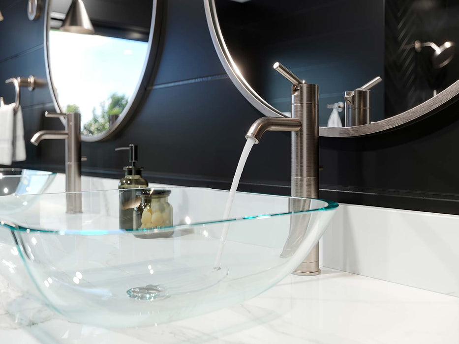 Pfister Contempra single-control vessel bathroom faucet in brushed nickel, installed on a white modern marble countertop, with water flowing into a glass basin and round brushed nickel edged mirrors in a modern bathroom.