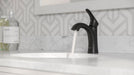 Pfister Weller single-control bathroom faucet in matte black, installed on a white countertop in a modern bathroom, with a steady stream of water flowing from the spout.