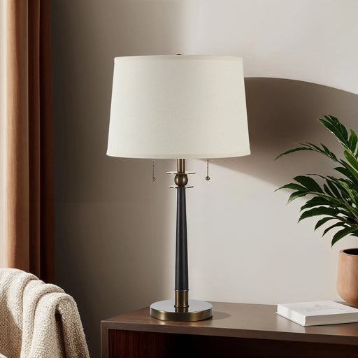 Table lamp with a white shade on a wooden side table next to a plant and books.