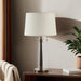 Table lamp with a white shade on a wooden side table next to a plant and books.