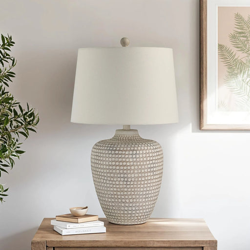 Textured table lamp on a wooden surface with a plant and framed artwork in the background.
