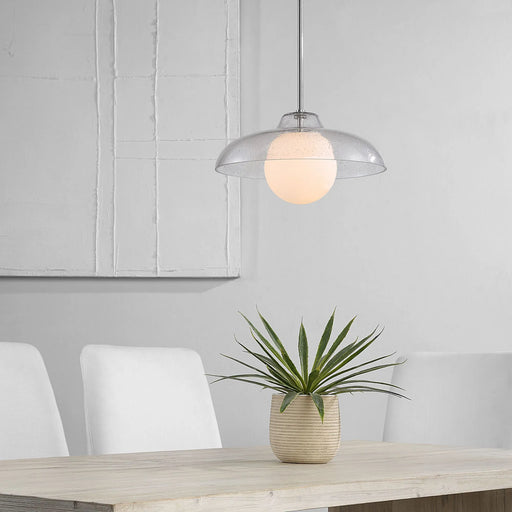 Clear pendant light fixture hanging above a dining table with a plant and chairs in the background.