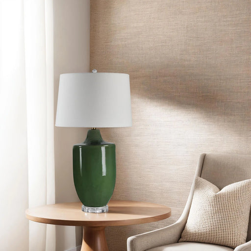 Green table lamp with white shade on a wooden side table against a textured wall.
