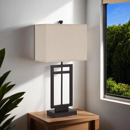 Table lamp with beige shade on a wooden table near a window with a view of greenery.