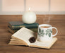 Ivory and green pinecone mug styled with an open book in a cozy setting