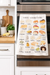 Cheese alphabet kitchen towel hanging on oven in modern kitchen setting with colorful cheese illustrations